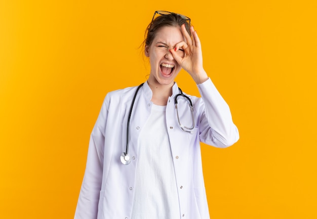 Jeune femme agacée en uniforme de médecin avec stéthoscope regardant à travers les doigts