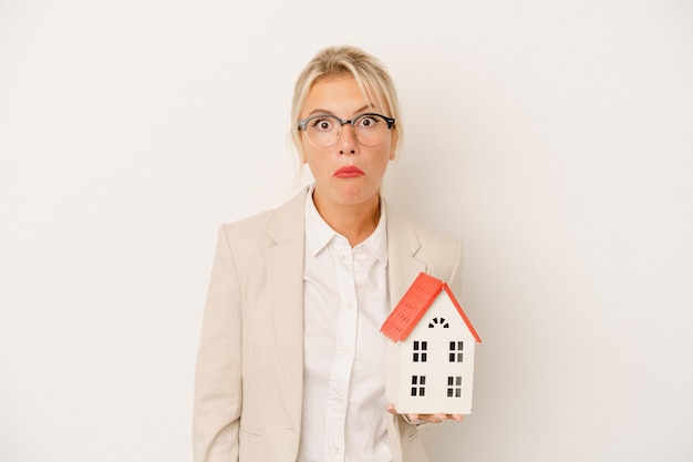 Jeune femme d'agent immobilier tenant un modèle de maison isolé sur fond blanc hausse les épaules et ouvre les yeux confus.