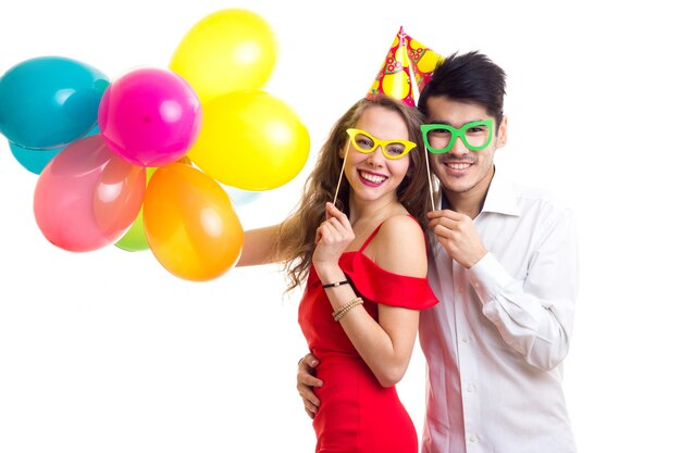 Jeune femme agréable aux longs cheveux bruns en robe rouge et jeune bel homme en chemise blanche avec des chapeaux de fête tenant de nombreux ballons colorés et des bâtons de lunettes sur fond blanc en studio