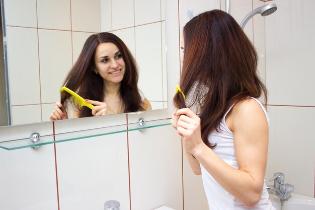 Jeune femme agréable se brossant les longs cheveux bruns avec un peigne devant le miroir de la salle de bain