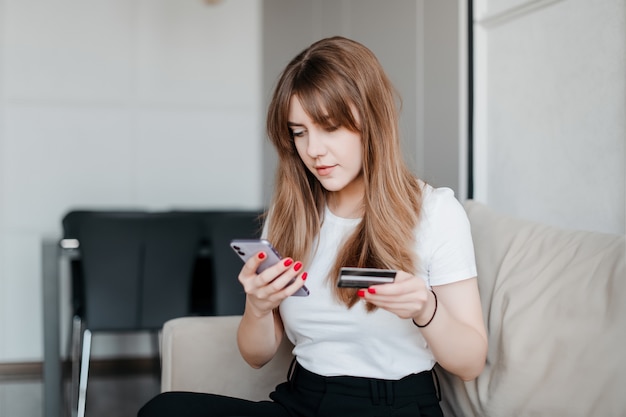 Jeune femme à l'aide de services bancaires mobiles avec carte de crédit et téléphone assis sur un canapé à l'appartement moderne