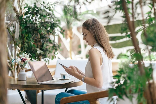 Jeune femme à l'aide d'un smartphone et d'un ordinateur portable assis à une table dans un café