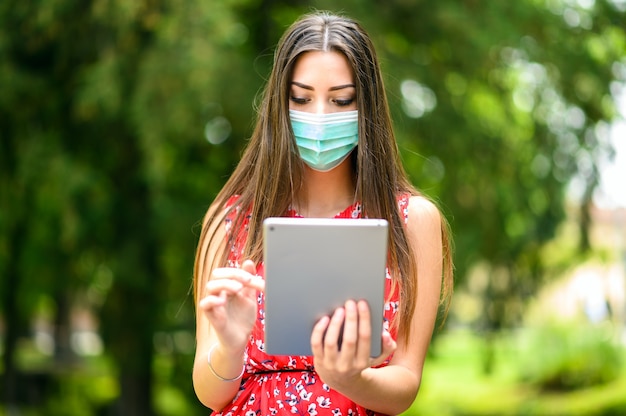 Jeune femme à l'aide d'une tablette numérique assise sur un banc dans un parc et portant un masque, concept de coronavirus