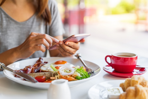 Jeune femme à l'aide de téléphone intelligent avec une tasse de café tout en mangeant le petit déjeuner