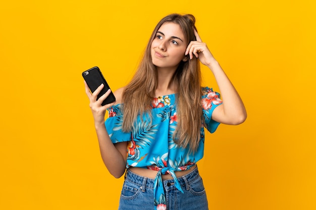 Jeune femme à l'aide de téléphone mobile sur mur jaune isolé ayant des doutes et avec l'expression du visage confus