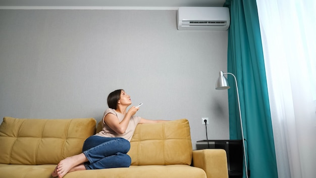 Jeune femme ajustant le climatiseur alors qu'elle était assise sur le canapé.