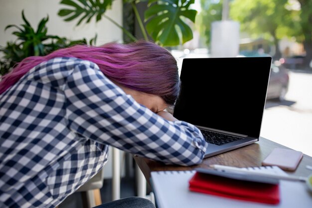 Jeune femme allongée sur les bras sur la table au café devant un ordinateur portable avec une tasse de café endormi