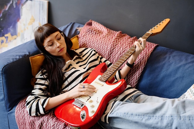 Jeune femme allongée sur un canapé et jouant de la guitare pendant son temps libre à la maison