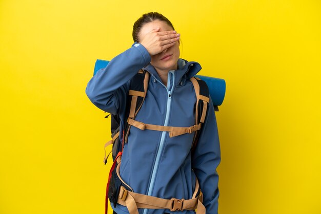 Jeune femme alpiniste avec un gros sac à dos sur fond jaune isolé couvrant les yeux à la main. Je ne veux pas voir quelque chose