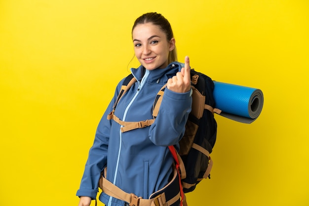 Jeune femme alpiniste avec un gros sac à dos sur fond jaune isolé faisant un geste à venir