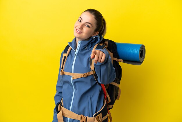 Jeune femme alpiniste avec un gros sac à dos sur fond jaune isolé pointant vers l'avant avec une expression heureuse