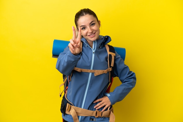 Jeune femme alpiniste avec un gros sac à dos sur fond jaune isolé souriant et montrant le signe de la victoire