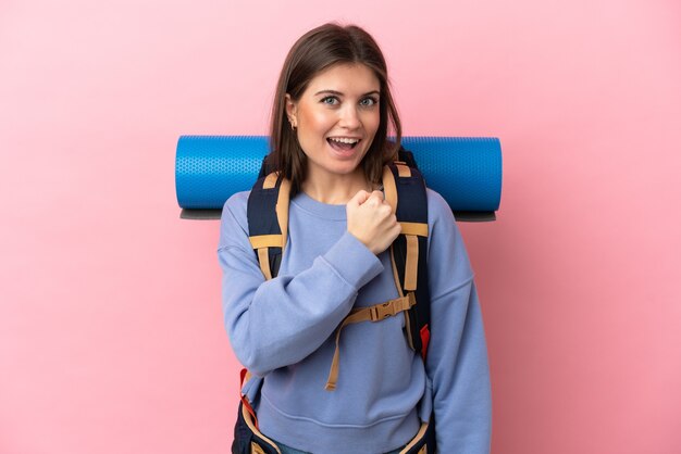 Jeune femme alpiniste avec un gros sac à dos isolé sur fond rose célébrant une victoire