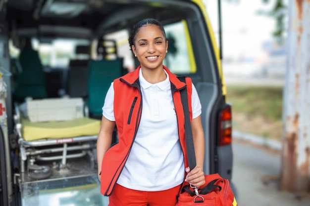 Jeune femme ambulancière debout à l'arrière d'une ambulance près des portes ouvertes Elle regarde la caméra avec une expression confiante souriant portant un sac de traumatologie médicale sur son épaule