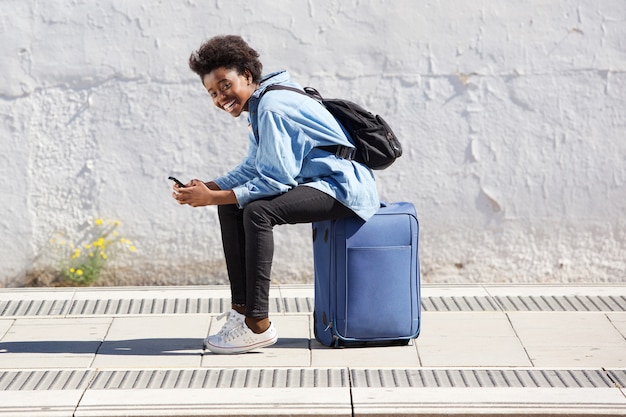 Jeune femme américaine afro assis sur une valise à la plate-forme de chemin de fer avec téléphone portable