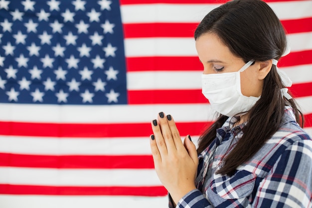 Jeune femme américaine avec le drapeau des USA en arrière-plan. Porter un masque contre le coronavirus et prier.