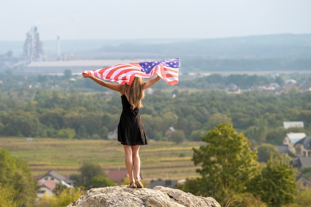 Jeune femme américaine heureuse aux cheveux longs se levant en agitant le vent drapeau national des États-Unis dans ses mains se relaxant à l'extérieur en profitant d'une chaude journée d'été.