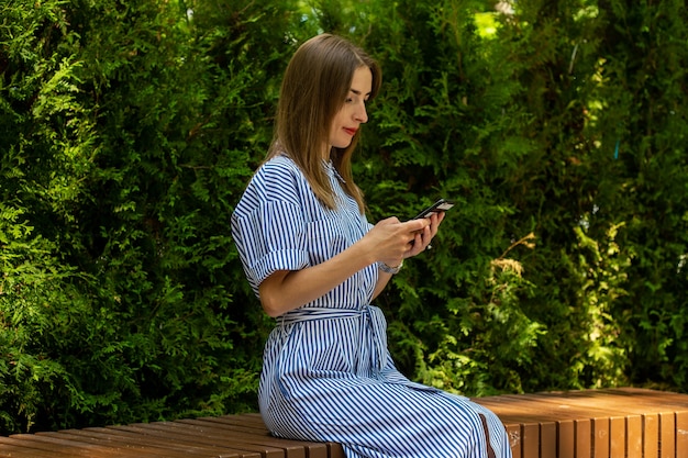 Jeune femme amicale dans une robe est assise sur un banc et regarde son téléphone dans le parc.