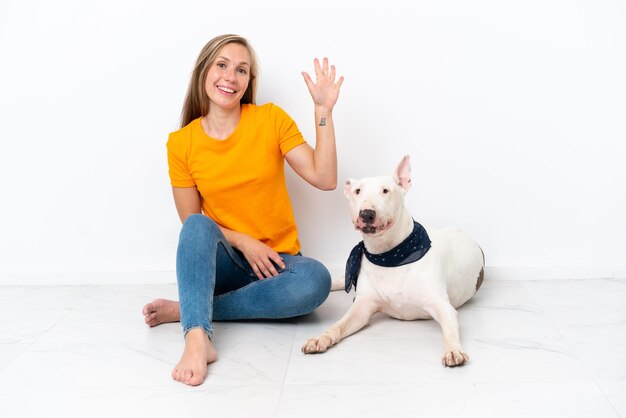 Jeune femme anglaise assise sur le sol avec son chien isolé sur fond blanc saluant avec la main avec une expression heureuse