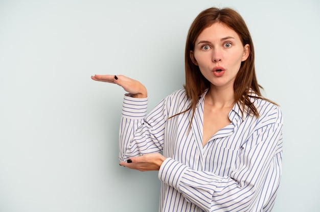 Jeune femme anglaise isolée sur fond bleu choquée et étonnée tenant un espace de copie entre les mains.