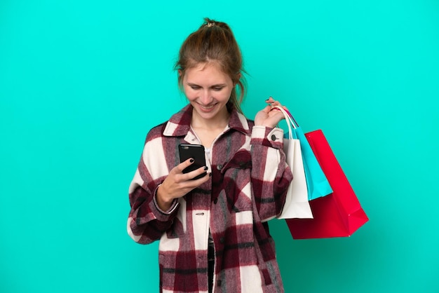 Jeune femme anglaise isolée sur fond bleu tenant des sacs à provisions et écrivant un message avec son téléphone portable à un ami