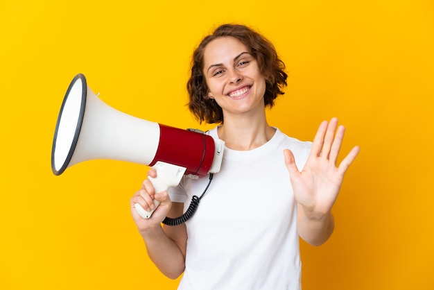 Jeune femme anglaise isolée sur fond jaune tenant un mégaphone et saluant avec la main avec une expression heureuse