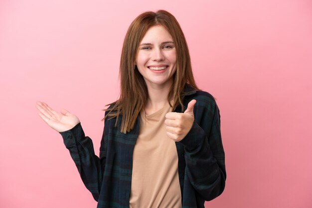 Jeune femme anglaise isolée sur fond rose tenant un fond imaginaire sur la paume pour insérer une annonce et avec les pouces vers le haut