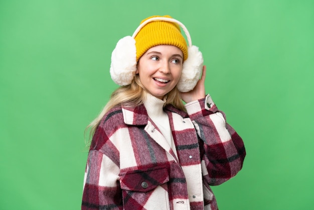 Jeune femme anglaise portant des manchons d'hiver sur fond isolé écoutant quelque chose en mettant la main sur l'oreille