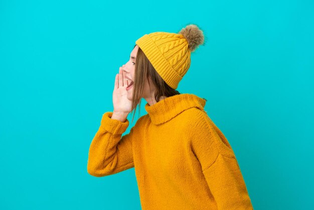 Jeune femme anglaise portant une veste d'hiver isolée sur fond bleu criant avec la bouche grande ouverte sur le côté