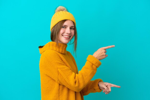 Jeune femme anglaise portant une veste d'hiver isolée sur fond bleu pointant le doigt sur le côté et présentant un produit