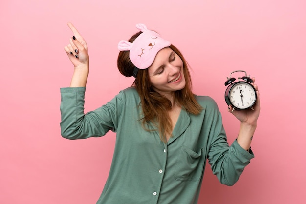 Jeune femme anglaise en pyjama isolée sur fond rose en pyjama et tenant une horloge avec une expression heureuse