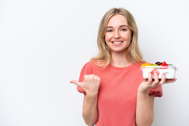 Jeune femme anglaise tenant un bol de fruits sur fond blanc isolé pointant vers le côté pour présenter un produit