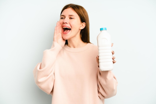 Jeune femme anglaise tenant une bouteille de lait isolée sur fond bleu criant et tenant la paume près de la bouche ouverte