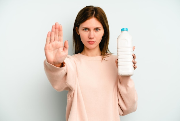 Jeune femme anglaise tenant une bouteille de lait isolée sur fond bleu debout avec la main tendue montrant le panneau d'arrêt, vous empêchant.