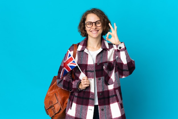 Jeune femme anglaise tenant un drapeau du Royaume-Uni isolé sur l'espace bleu montrant signe ok avec les doigts