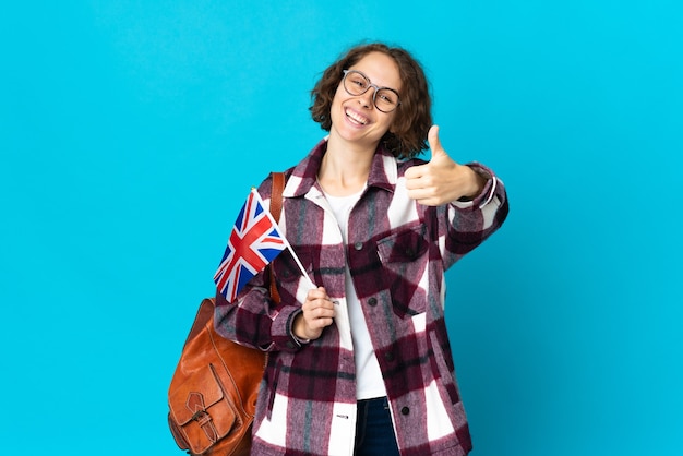 Jeune femme anglaise tenant un drapeau du Royaume-Uni isolé sur un mur bleu avec les pouces vers le haut parce que quelque chose de bien s'est passé