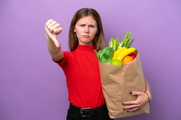 Jeune femme anglaise tenant un sac d'épicerie isolé sur fond violet montrant le pouce vers le bas avec une expression négative