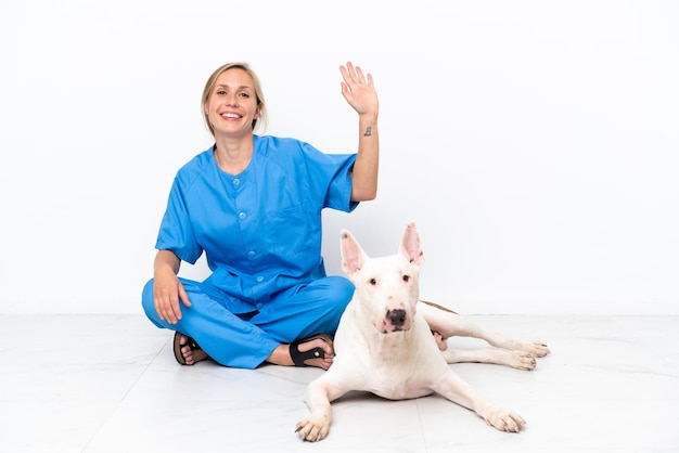 Jeune femme anglaise vétérinaire assise sur le sol avec un chien saluant avec la main avec une expression heureuse