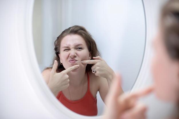 Une jeune femme anxieuse se regarde dans le miroir, inquiète des rides ou de l'acné sur une peau malsaine, contrariée par une femme du millénaire malheureuse, examine le bouton pressé sur le visage dans la chambre à coucher concept de soins de la peau en cosmétologie