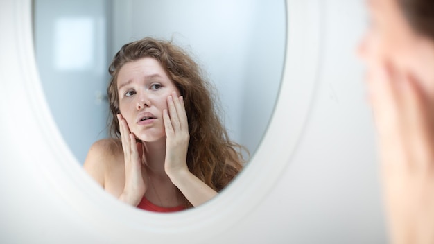 Une jeune femme anxieuse se regarde dans le miroir, inquiète des rides ou de l'acné sur une peau malsaine, contrariée par une femme du millénaire malheureuse, examine le bouton pressé sur le visage dans la chambre à coucher concept de soins de la peau en cosmétologie