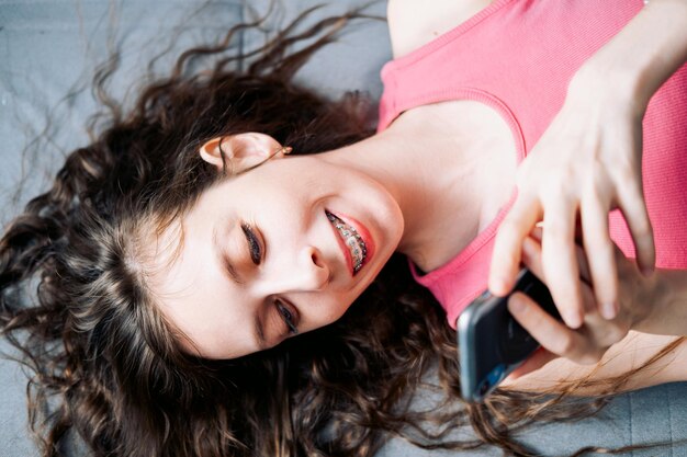 Une jeune femme en appareil orthopédique avec de longs cheveux bouclés est allongée sur le canapé et parcourt les réseaux sociaux sur son téléphone.