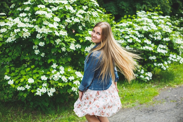 Une jeune femme d'apparence européenne aux longs cheveux blonds, vêtue d'une robe courte, se dresse sur fond de buissons à fleurs blanches. Journée de printemps ensoleillée. Beauté féminine naturelle