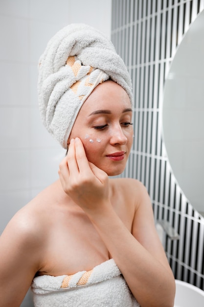 Jeune femme appliquant une crème anti-rides debout derrière un miroir dans la salle de bain à domicile Procédure de cosmétologie et de beauté Soins de la peau après le nettoyage