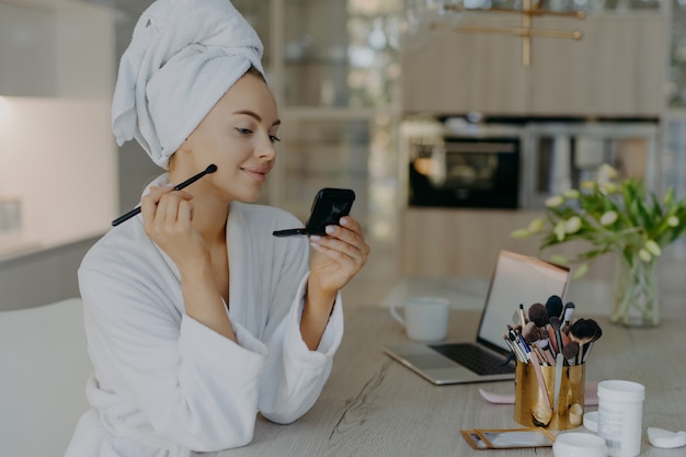 jeune femme applique des cosmétiques de maquillage avec une brosse en face du miroir