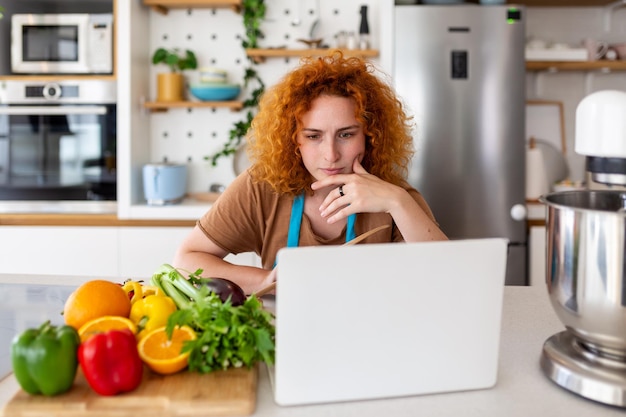 Une jeune femme apprend à cuisiner, elle regarde des recettes vidéo sur un ordinateur portable dans la cuisine et cuisine un plat Concept de cuisine à la maison