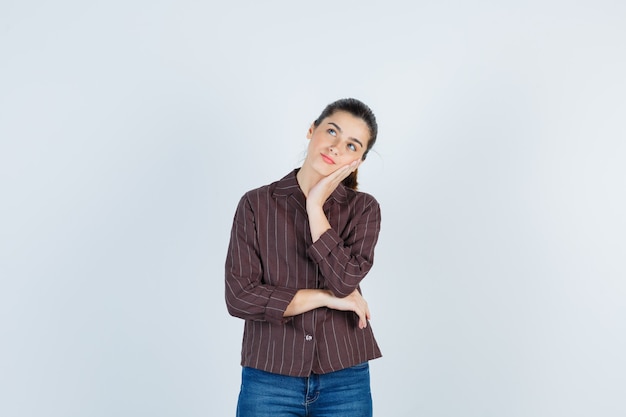 Jeune femme appuyée sur la joue de la paume, pensant à quelque chose en chemise rayée, jeans et regardant pensive, vue de face.