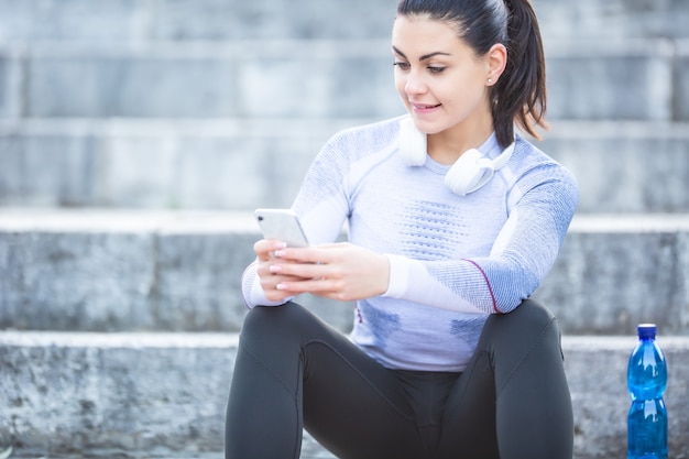 La jeune femme après la formation vérifie le tracker de sport. Concept de remise en forme, casque de sous-vêtements thermiques et smartphone.