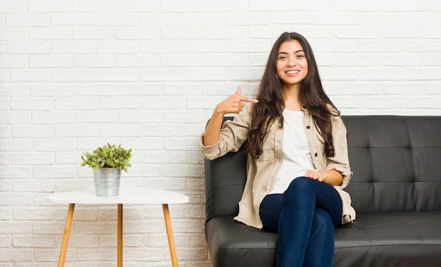 Jeune femme arabe assise sur le canapé personne pointant à la main vers un espace copie de chemise, fier et confiant