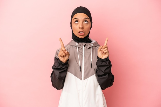 Jeune femme arabe avec burqa de sport isolée sur fond rose pointant vers le haut avec la bouche ouverte