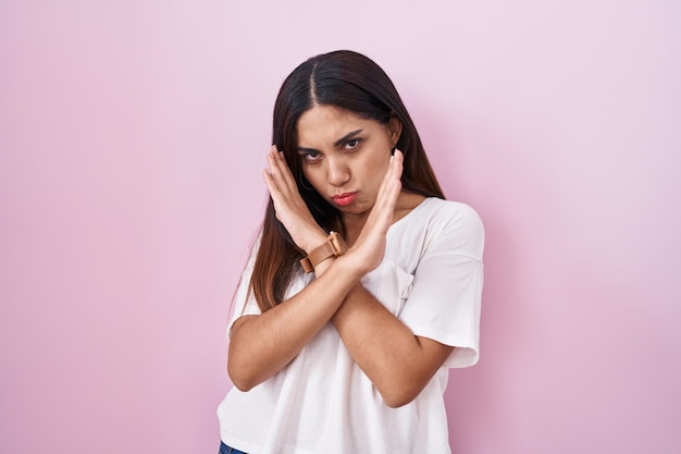 Jeune femme arabe debout sur fond rose expression de rejet croisant les bras faisant signe négatif, visage en colère